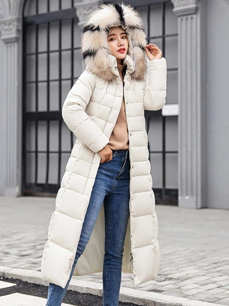Femme portant un manteau long en duvet blanc avec capuche en fourrure, posant devant un bâtiment urbain. Mode hiver, style élégant, tenue chaude.