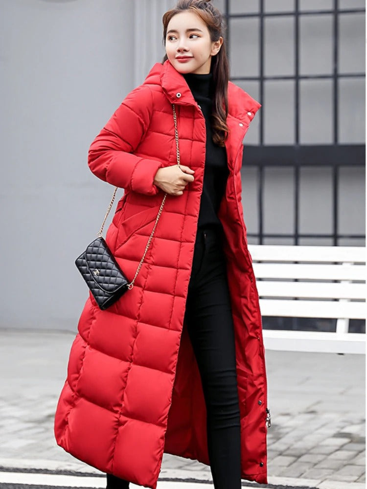 Femme portant un long manteau d'hiver rouge, sac à main noir, posant devant un banc blanc. Mode féminine, tenue chaude, style urbain.
