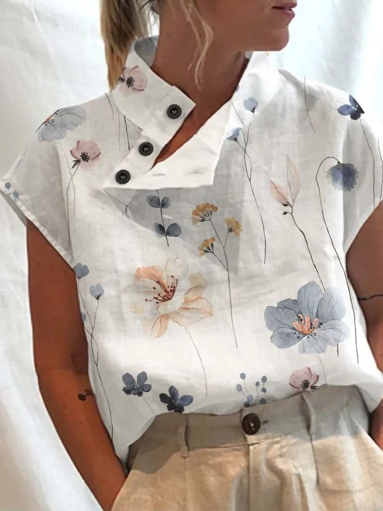 Femme portant une chemise blanche à motifs floraux bleus et beiges, col boutonné, manches courtes, associée à un pantalon beige. Mode élégante et décontractée.