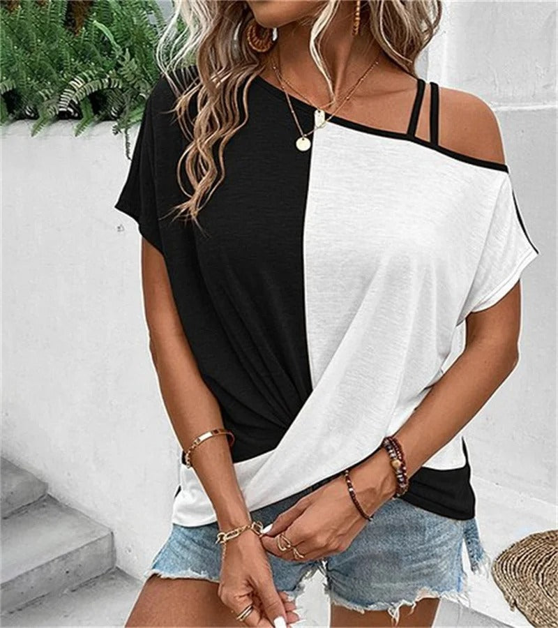Femme portant un t-shirt bicolore noir et blanc, épaules dénudées, avec des bijoux dorés, shorts en jean, posant devant un mur blanc et des plantes.