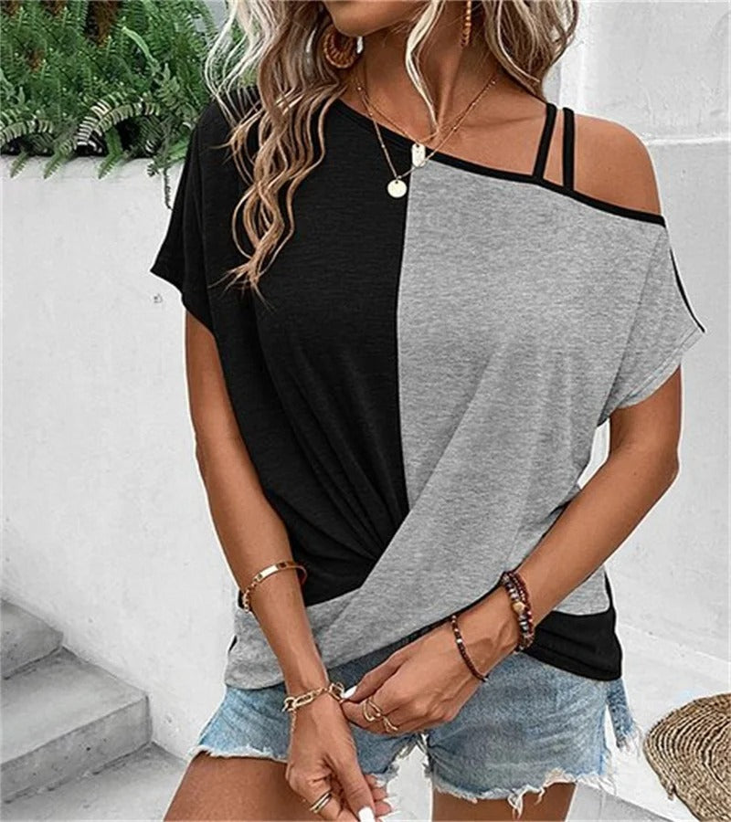 Femme portant un t-shirt bicolore noir et gris, épaules dénudées, avec des bijoux dorés, shorts en jean, posant devant des plantes vertes.