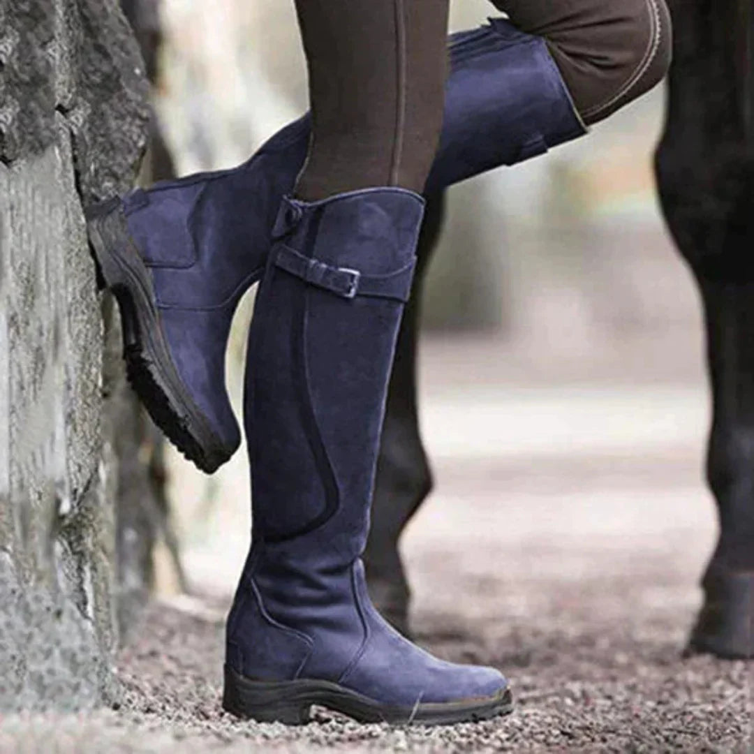 Bottes d'équitation en cuir bleu marine, portées par une personne appuyée contre un mur, sur fond de sol en gravier. Accessoire équestre élégant.
