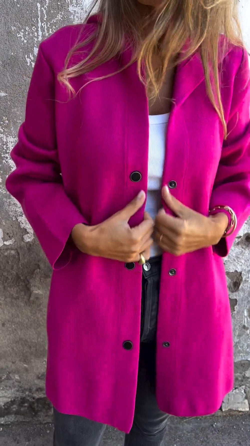 Femme portant un manteau fuchsia élégant, boutons noirs, sur un mur en pierre. Mode automne, style chic, tendance vestimentaire féminine.