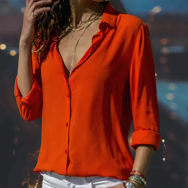 Femme portant une chemise orange vif à manches longues, col ouvert, avec des accessoires dorés, posant devant un fond flou. Mode élégante et moderne.