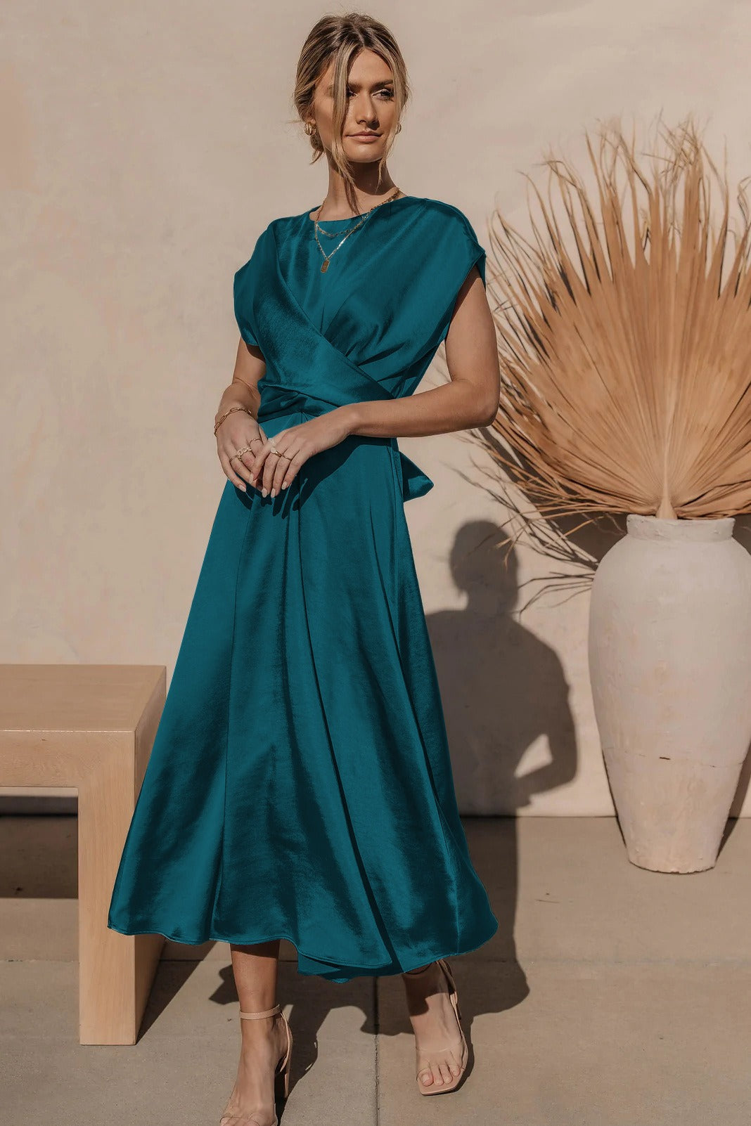 Robe longue en satin bleu-vert, femme debout près d'un vase en céramique blanche avec feuilles séchées, style élégant et moderne, mode féminine.