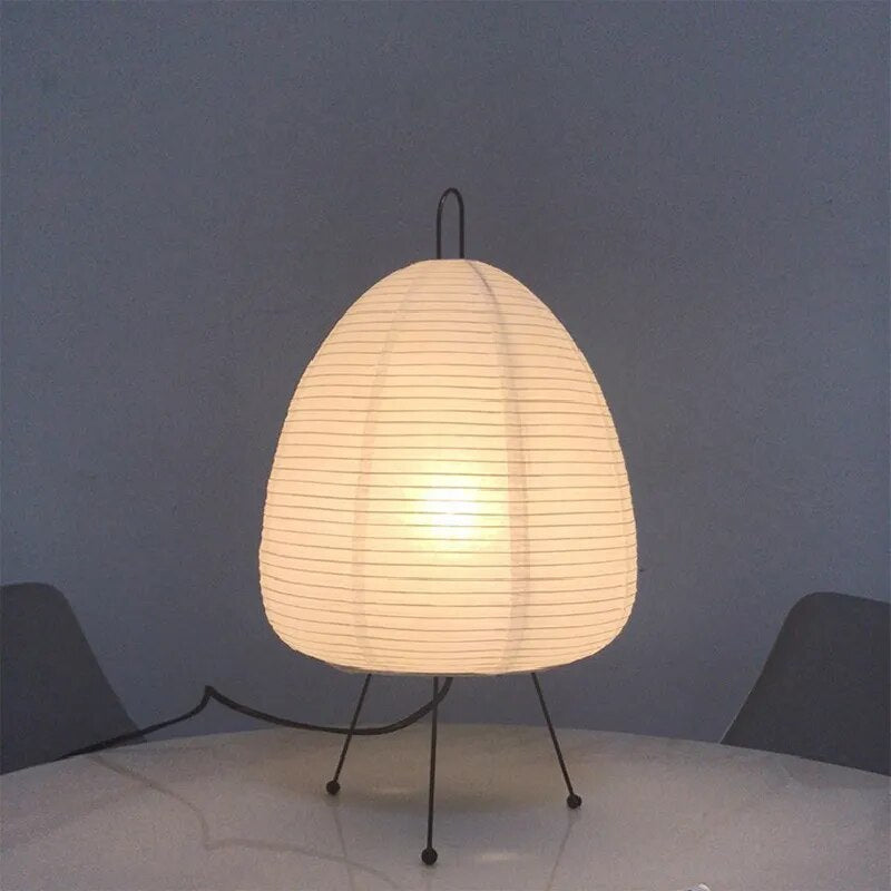 Lampe de table en papier blanc sur trépied noir, éclairée, posée sur une table blanche. Design moderne, éclairage intérieur élégant.