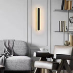 Applique murale LED moderne noire et dorée éclairée, décor minimaliste avec fauteuils gris et étagère design, salon contemporain élégant.
