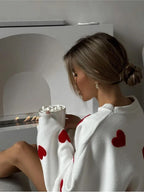 Femme buvant un chocolat chaud, pull blanc à cœurs rouges, devant une cheminée moderne. Ambiance cosy, détente hivernale, style cocooning.