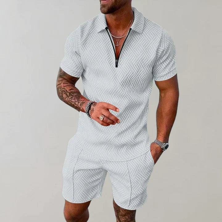 Homme portant un ensemble short et polo blanc à motifs géométriques, style décontracté, avec tatouages visibles sur le bras, fond neutre.