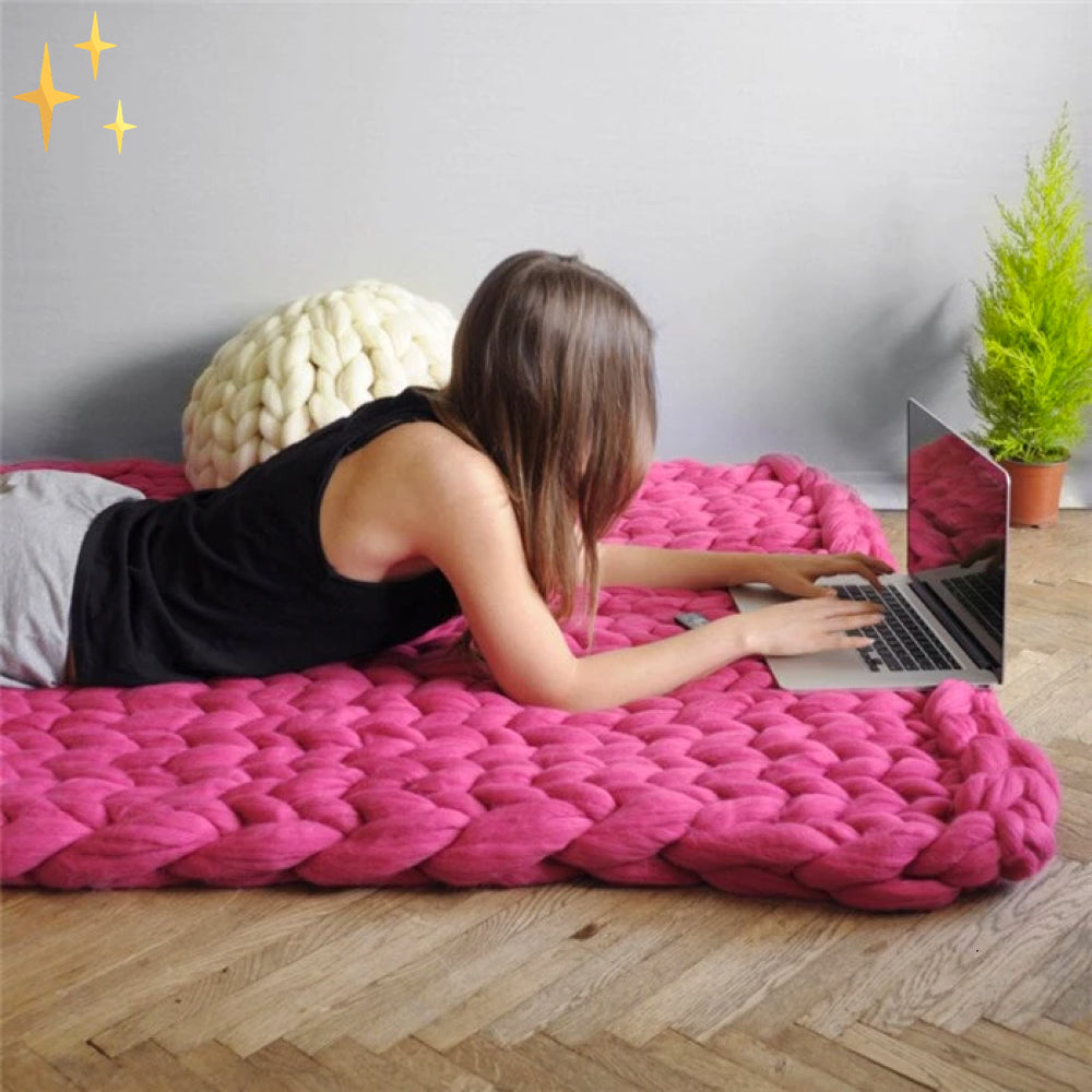 Femme allongée sur un tapis en laine rose tricoté, utilisant un ordinateur portable. Décor moderne avec plante verte et coussin en arrière-plan.