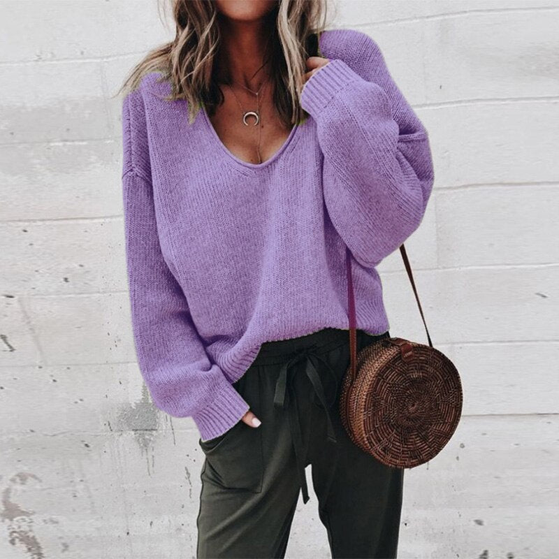 Femme portant un pull violet ample, pantalon vert foncé, sac rond en osier, posant devant un mur blanc. Mode décontractée, tendance automne.