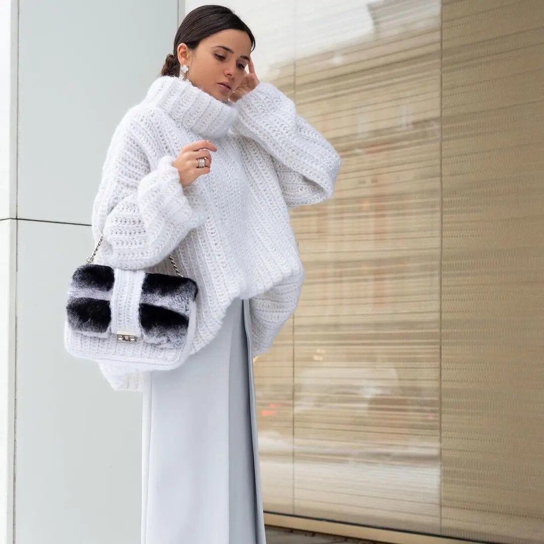 Femme portant un pull en maille blanc, jupe longue grise, sac à main en fourrure noir et blanc, posant devant un mur moderne. Mode hiver élégante.