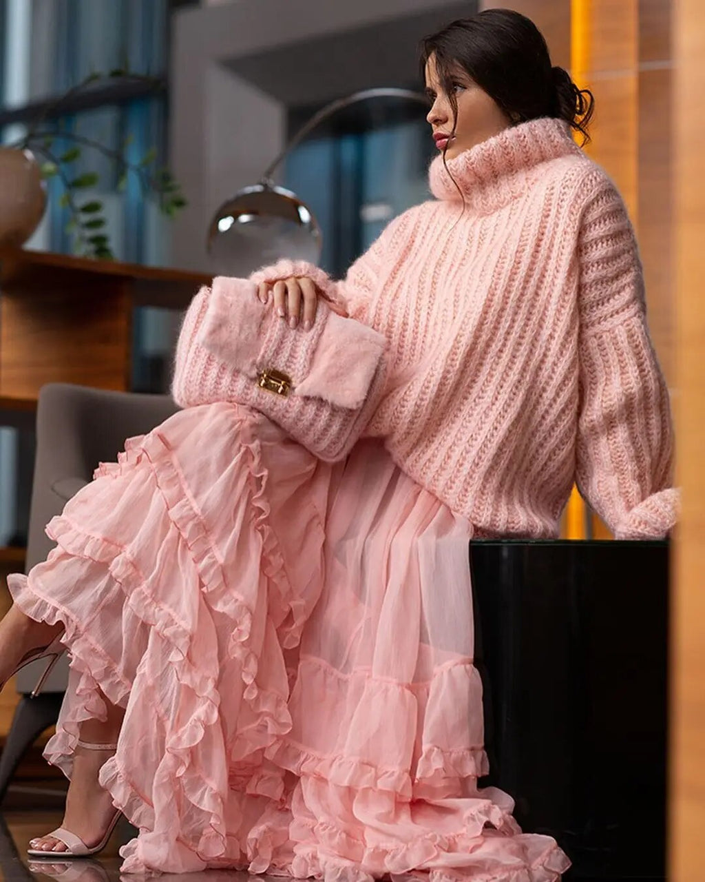 Femme en pull rose épais et jupe à volants, assise dans un intérieur moderne. Mode automne-hiver, style élégant et confortable.