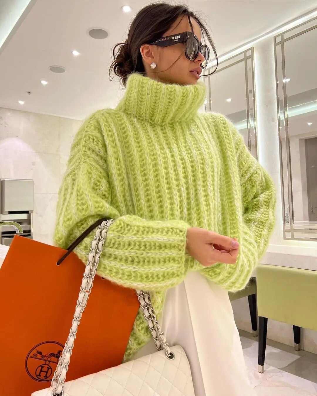 Femme portant un pull vert en maille épaisse, lunettes de soleil, sac orange et sac blanc à chaîne, dans un intérieur moderne et lumineux.