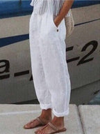 Personne portant un pantalon blanc en lin, sandales marron, debout près d'un bateau. Mode estivale, tenue décontractée, bord de mer.