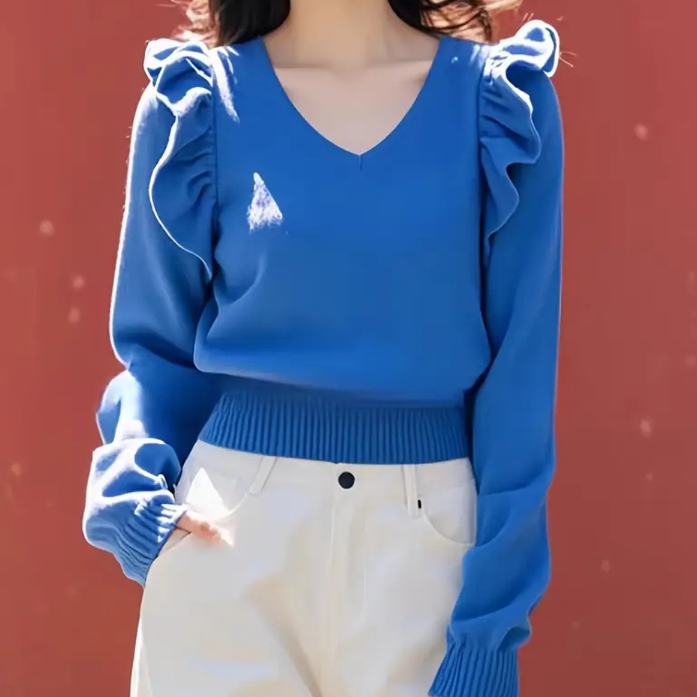 Femme portant un pull bleu à volants avec un col en V, manches longues, associé à un pantalon blanc. Mode féminine élégante et moderne.