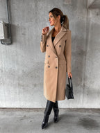 Femme élégante portant un manteau beige, bottes noires et sac à main, posant contre un mur gris. Mode automne chic, style urbain sophistiqué.
