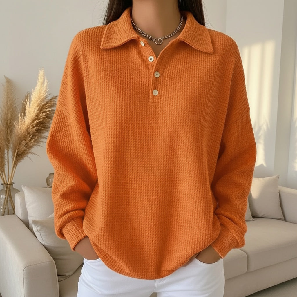 Pull orange à col polo et boutons, porté par une personne debout dans un salon moderne. Mode automne, style décontracté, intérieur lumineux.