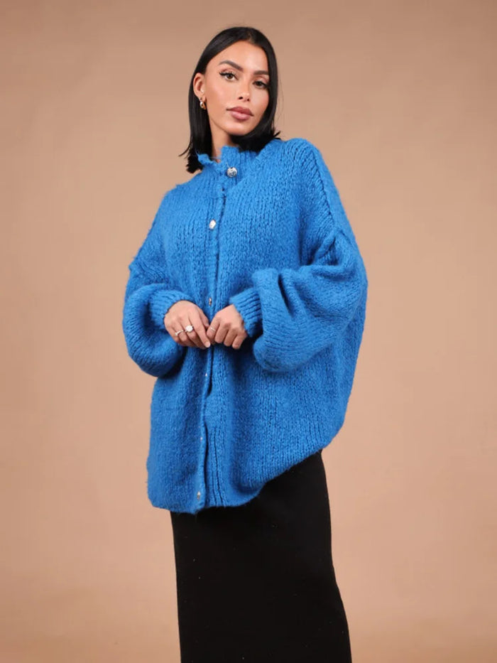 Femme portant un cardigan bleu en laine épaisse, manches bouffantes, sur fond beige. Mode automne-hiver, style décontracté et élégant.