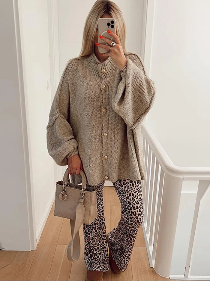 Femme en pull oversize beige, pantalon léopard, tenant un sac à main beige, se prenant en photo dans un miroir. Mode automne, style décontracté.