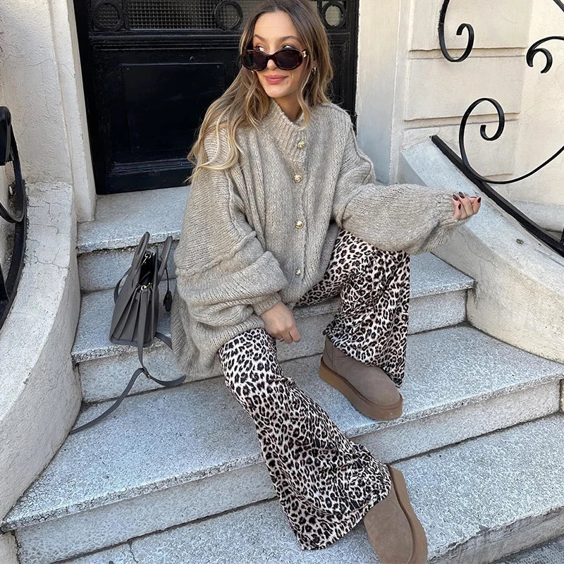 Femme assise sur des marches, portant un manteau gris ample, pantalon imprimé léopard, bottes marron et lunettes de soleil, avec un sac à main gris.