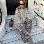 Femme assise sur des marches, portant un manteau gris ample, pantalon imprimé léopard, bottes marron et lunettes de soleil, avec un sac à main gris.