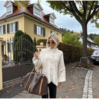 Femme élégante en pull beige devant maison traditionnelle, portant un grand sac de voyage marron. Arbre et voiture en arrière-plan.