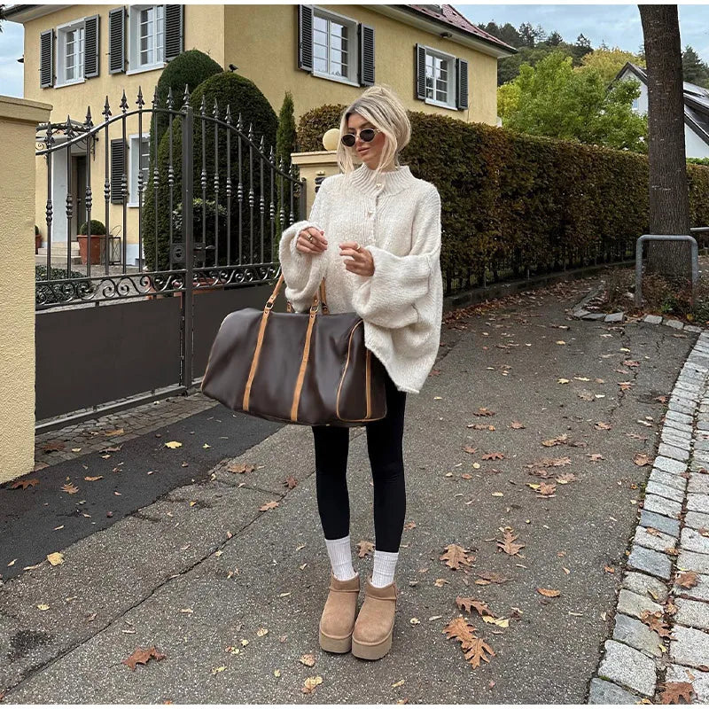 Femme en pull blanc oversize, leggings noirs, bottes beiges, portant un grand sac de voyage marron devant une maison jaune, automne, feuilles au sol.