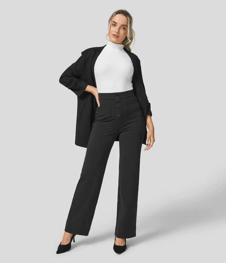 Femme en tenue professionnelle chic, portant un blazer noir, un col roulé blanc et un pantalon taille haute noir, posant sur fond gris.
