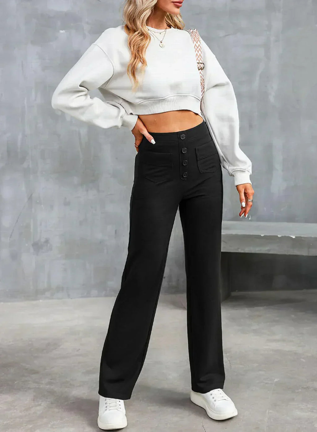 Femme portant un pull blanc court et un pantalon noir taille haute, posant contre un mur gris. Mode décontractée, style moderne, tenue élégante.