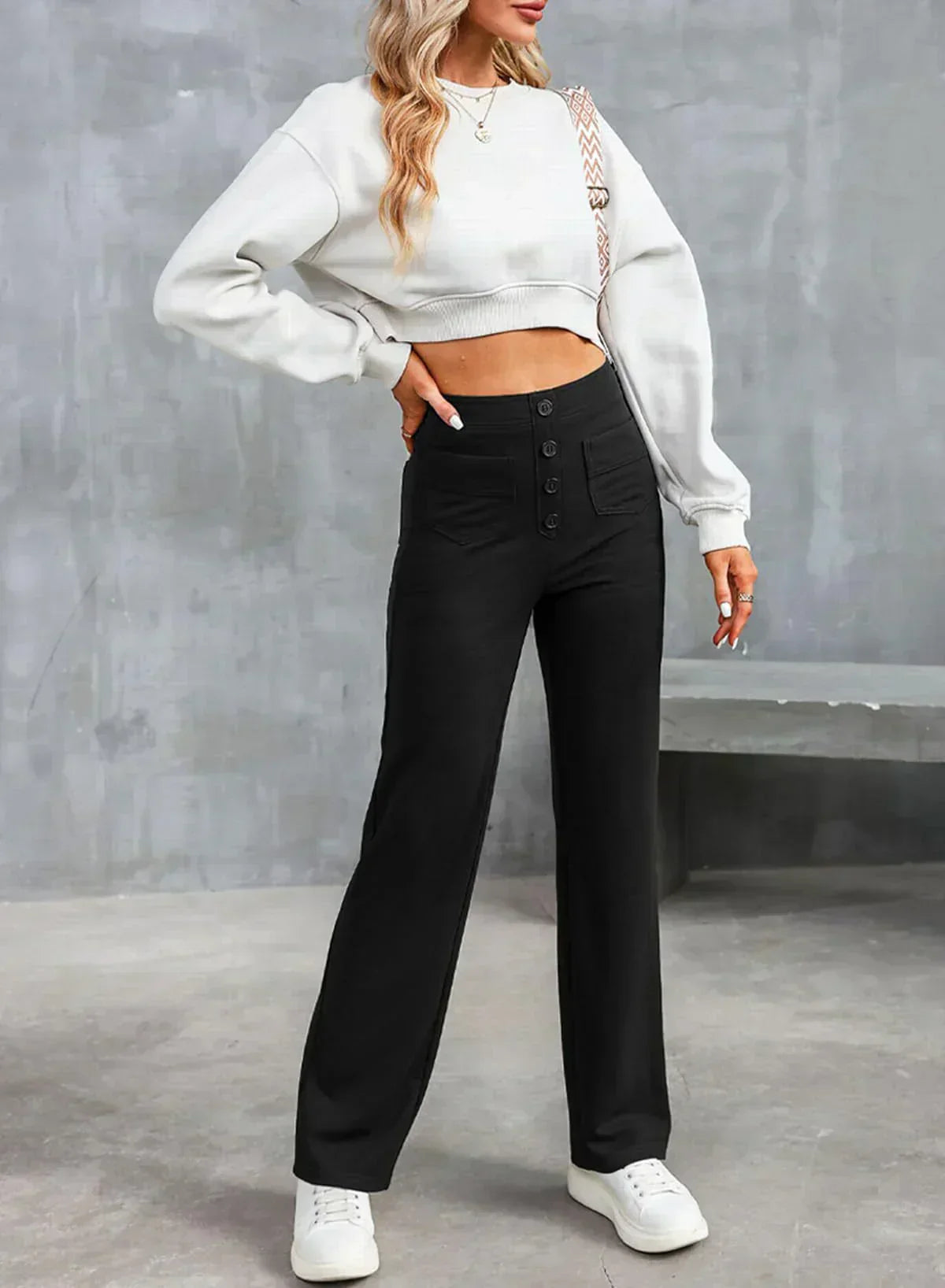 Femme portant un pull blanc court et un pantalon noir taille haute, posant contre un mur gris. Mode décontractée, style moderne, tenue élégante.