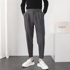 Homme en pantalon gris élégant, pull noir, baskets blanches, debout dans une pièce moderne. Mode masculine, style décontracté chic.