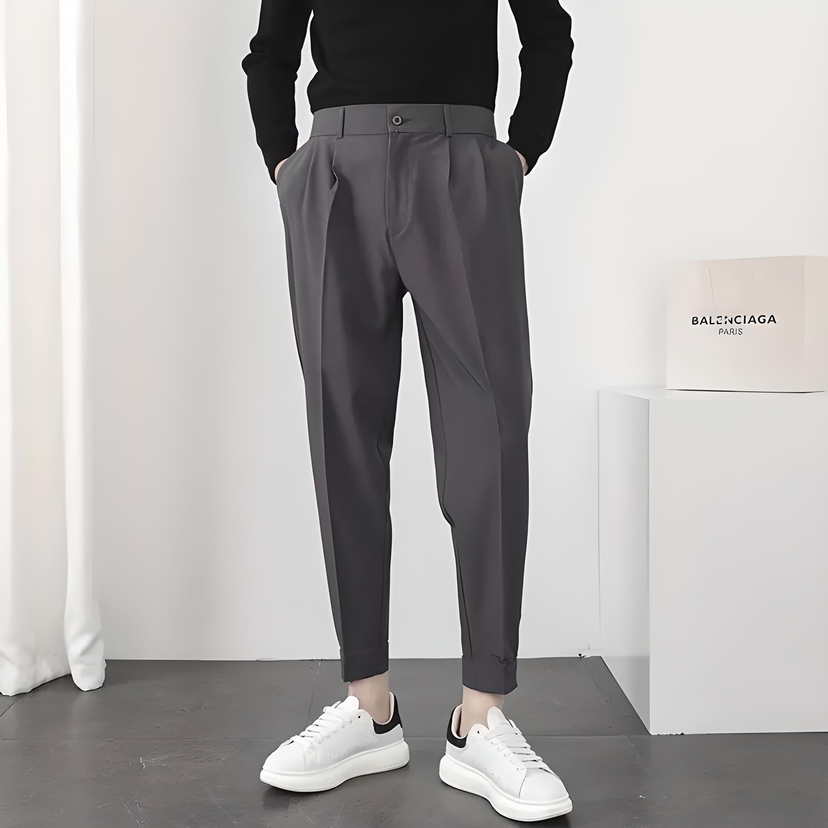 Homme en pantalon gris élégant, pull noir, baskets blanches, debout dans une pièce moderne. Mode masculine, style décontracté chic.